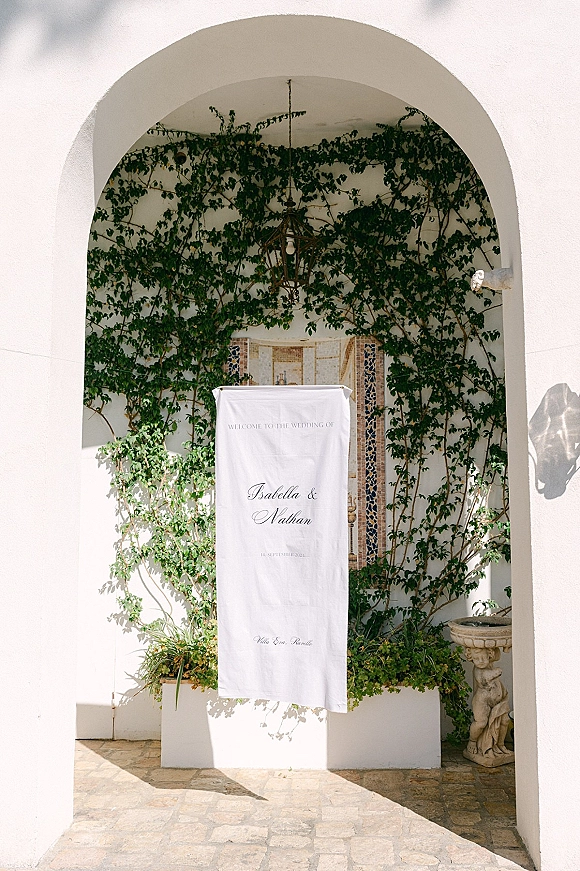 Wedding welcome sign fabric banner with calligraphy text hanging from a white stucco archway, framed by ivy and a mosaic tile fountain