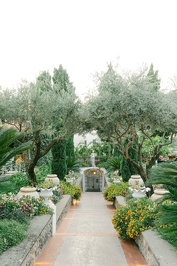 Garden wedding walkway lined with stone planters and potted flowers, with pathway lights leading to an arched gate amid olive trees