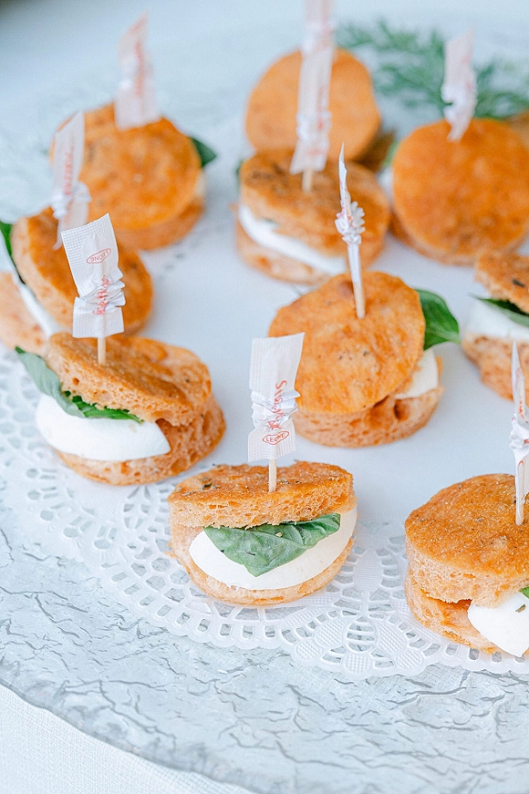 Wedding appetizers arranged as mini sandwich bites with cheese and basil, secured by flag toothpicks on a lace doily platter on white tabletop