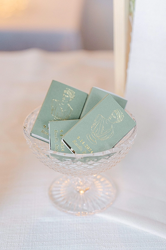 Wedding matchbooks with gold foil stamping in a crystal glass bowl, green covers displayed on a white tablecloth by a mirror frame