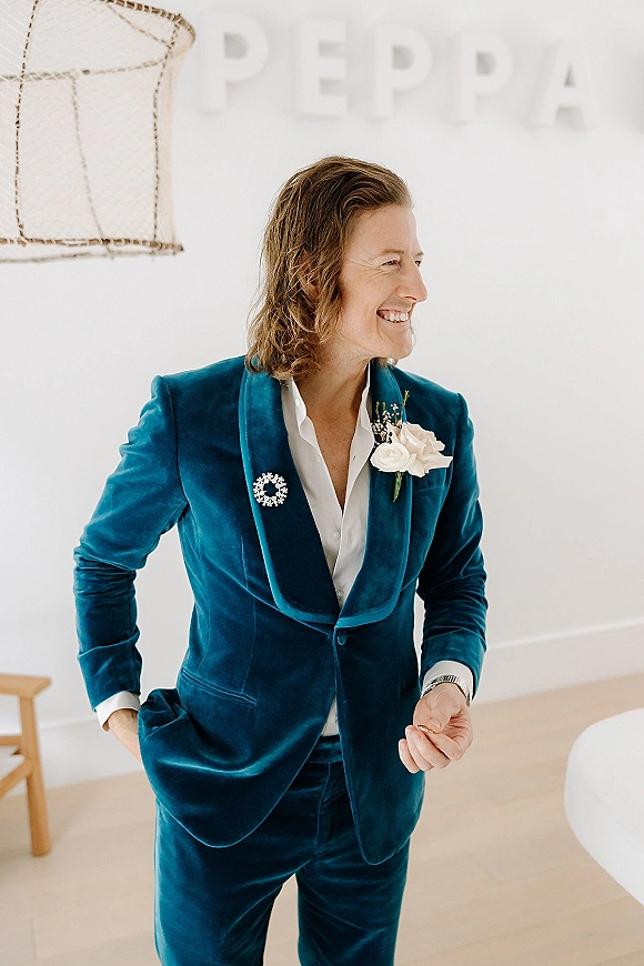Groom portrait in a velvet wedding suit with a white rose boutonniere, standing by a white wall with letters and hanging light fixture