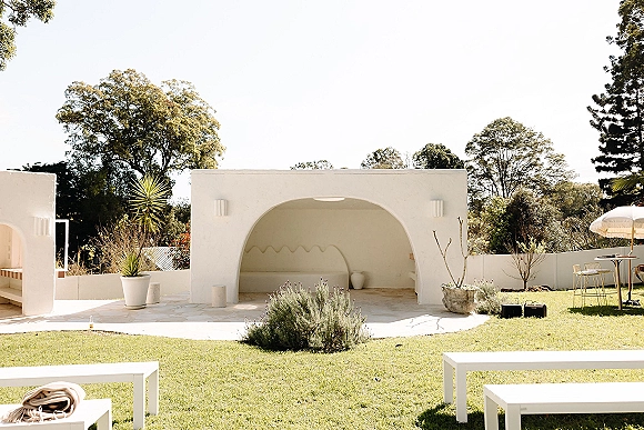 Outdoor lounge setup with white benches and patio umbrella, styled as a wedding lounge area with bar stools, plants, and lanterns on a lawn