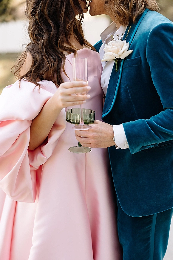 Wedding kiss portrait of newlyweds champagne toast, kissing while holding flutes as the groom wears a teal velvet suit outdoors