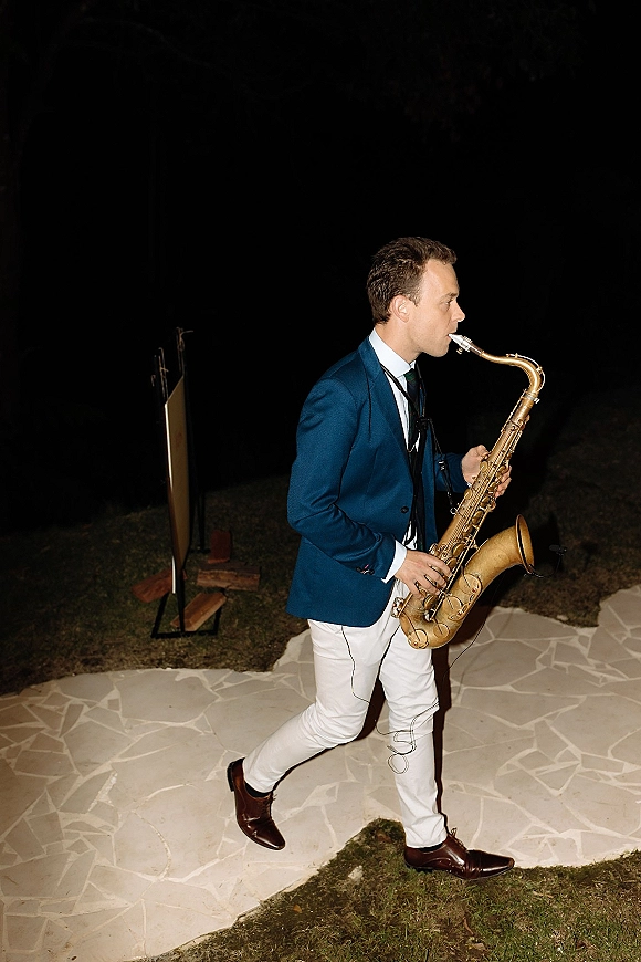 Wedding saxophonist, a live wedding musician, walks while playing with a saxophone accent on a stone pathway under the night sky