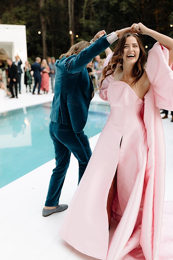 Wedding reception dancing as bride in a pink statement gown twirls with groom in a blue velvet suit on a poolside patio under string lights