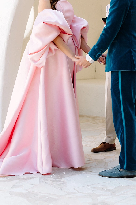 Ceremony moment as couple holding hands, bride in pink puff-sleeve gown and groom in blue suit against a white stucco wall backdrop