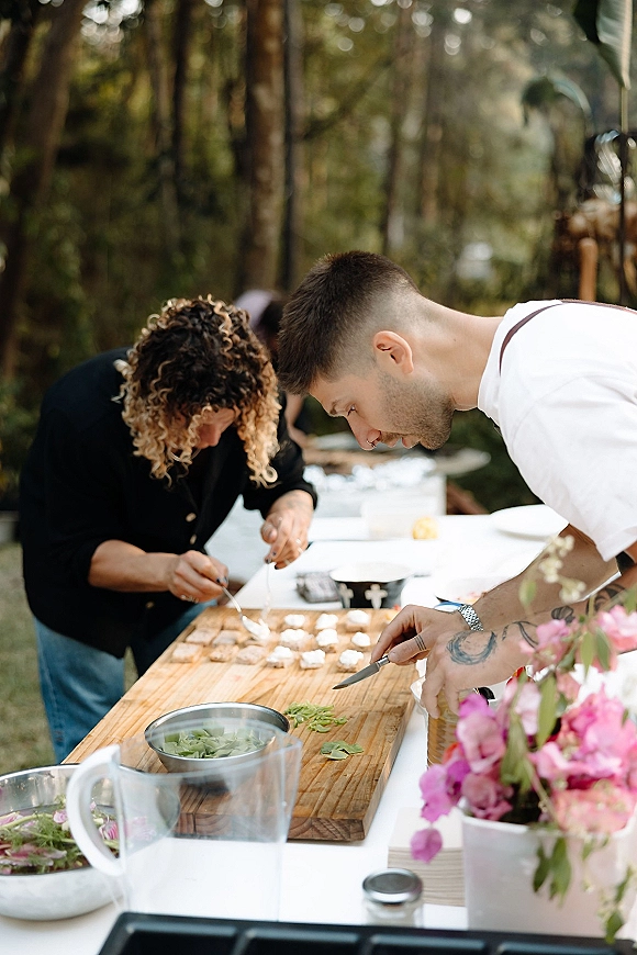 Wedding catering prep with a chef plating appetizers on a wood cutting board, herbs and edible flowers on a white tablecloth outdoors