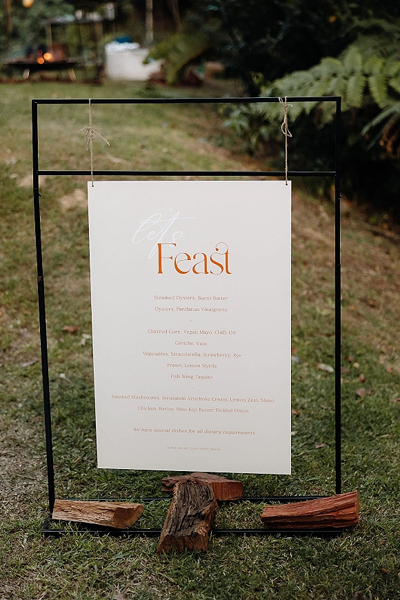 Wedding menu sign on a black metal sign frame, white poster tied with twine, set on logs amid ferns by a fire pit lawn