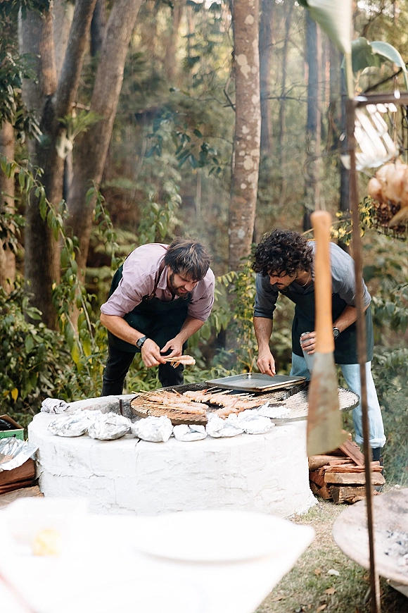 Wedding catering with shrimp skewers grilling over a live fire, chef in apron tending charcoal grill in a forest setting