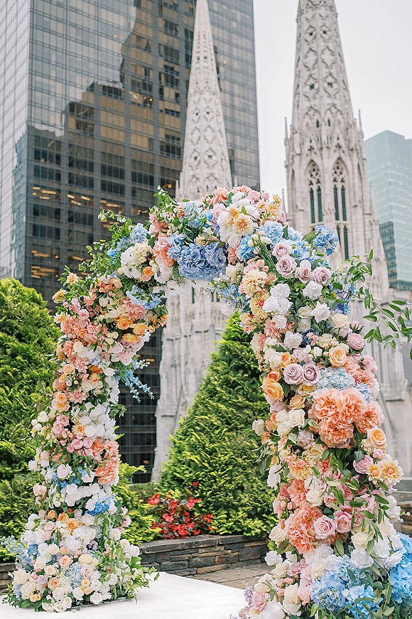 Wedding ceremony arch with a floral wedding arch of pastel roses and hydrangea, set on a rooftop terrace with city skyline and church spires behind