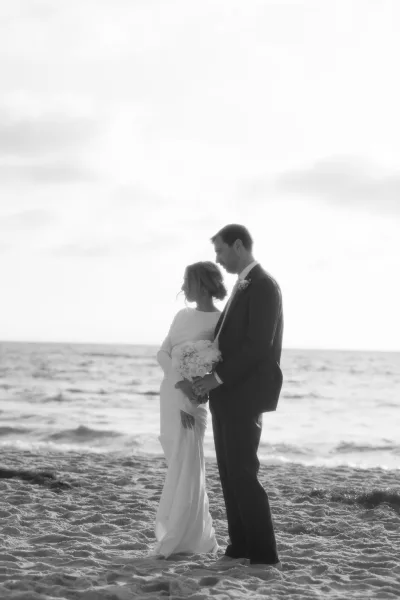 Couple portrait on the shoreline, a serene beach wedding portrait as the bride lifts a simple white bouquet with ocean waves and sky beyond.