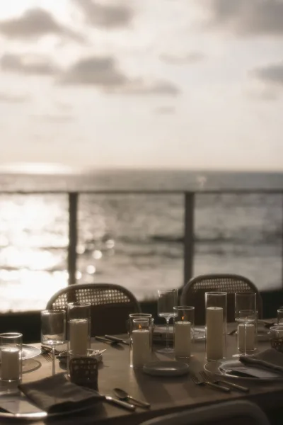 Reception tablescape sets a candlelit wedding table with pillar candles, framed by an ocean view and sunset sky beyond a balcony railing.