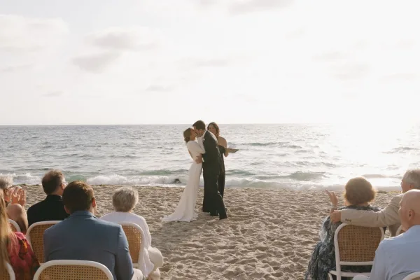 Ceremony kiss moment at a beach wedding ceremony, the couple embracing, the bride in a bridal gown against a sunlit ocean and sandy shore.