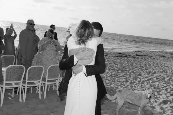 Ceremony moment as a bride and groom hug on a beach, ocean behind, guests applaud while their dog waits by the aisle.