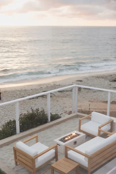Outdoor lounge setup offering beach lounge seating around a fire pit, set on the sand with ocean waves and a pastel sunset beyond.