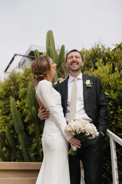 Couple portrait of the bride and groom laughing as she holds a bouquet, framed by cactus and hedges with a bright sky beyond.