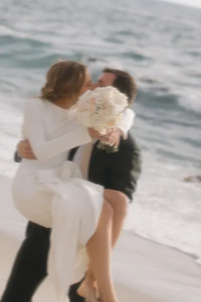 Wedding kiss portrait on a sandy beach with ocean waves, bride holding a white and blush bouquet as the groom leans in for a tender embrace.