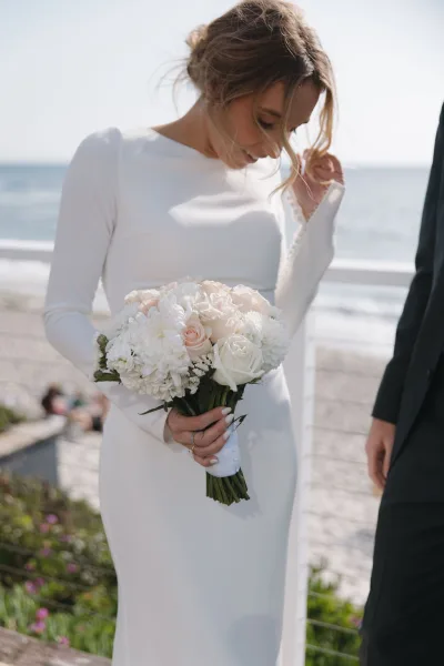 Bridal portrait of a bride holding bouquet, wearing a long-sleeve gown, set against an oceanfront beach with sky and coastal plants.