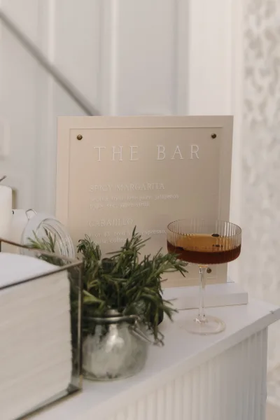 Wedding bar sign displaying a signature drink sign, with a cocktail glass nearby, set against a white wall and staircase.