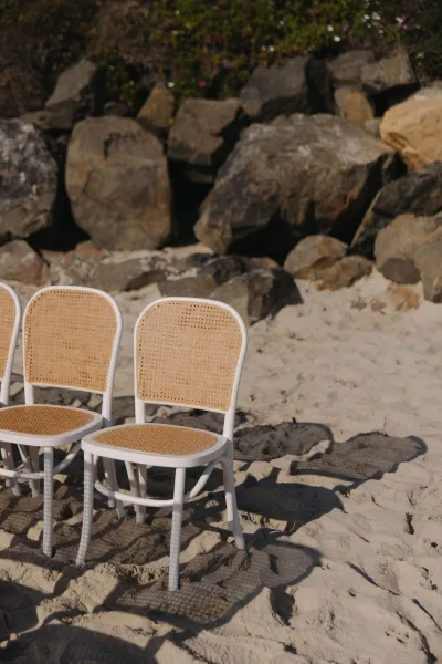 Beach ceremony seating with rattan wedding chairs in neat rows on a sand beach dotted with rocks, featuring cane webbing for airy texture.