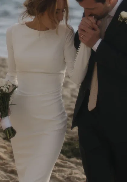 Couple portrait with the groom kissing the bride's hand, her wedding ring catching light, on beach sand with the ocean softly behind.