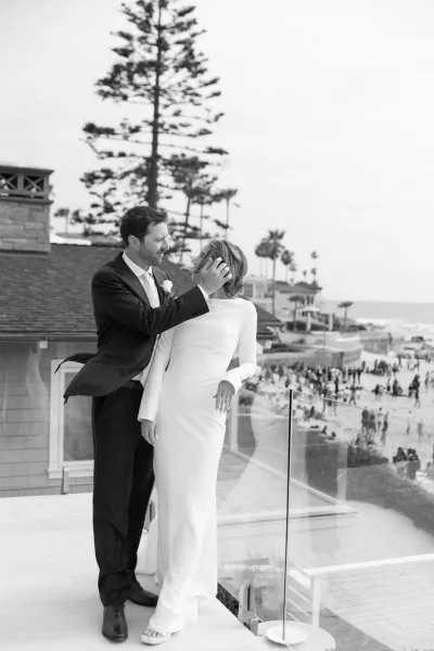 Couple portrait of a bride and groom embrace on a balcony above a beach and ocean, palm trees beyond, her wedding ring catching light in sea breeze.