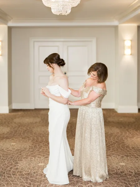 Bride getting ready as her mother helps button her off-the-shoulder gown, bride in updo with jewelry in a hotel hallway with chandelier