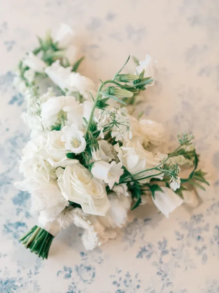 Bridal bouquet of white flowers with roses and tulips, wrapped in lace ribbon with greenery, resting on a blue floral patterned surface
