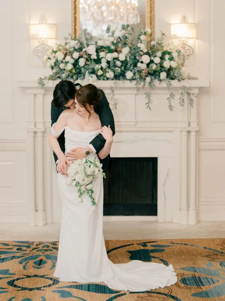 Couple portrait of bride and groom embrace by a white fireplace mantel, groom hugging from behind as she holds a cascading bouquet