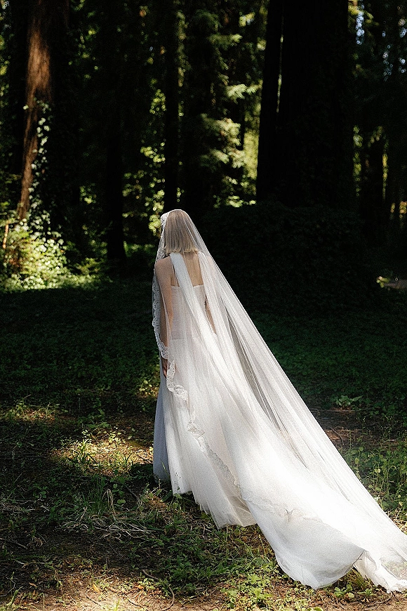 Bridal portrait of a bride from behind in a backless wedding dress with a long lace-trim veil flowing through a sunlit forest path