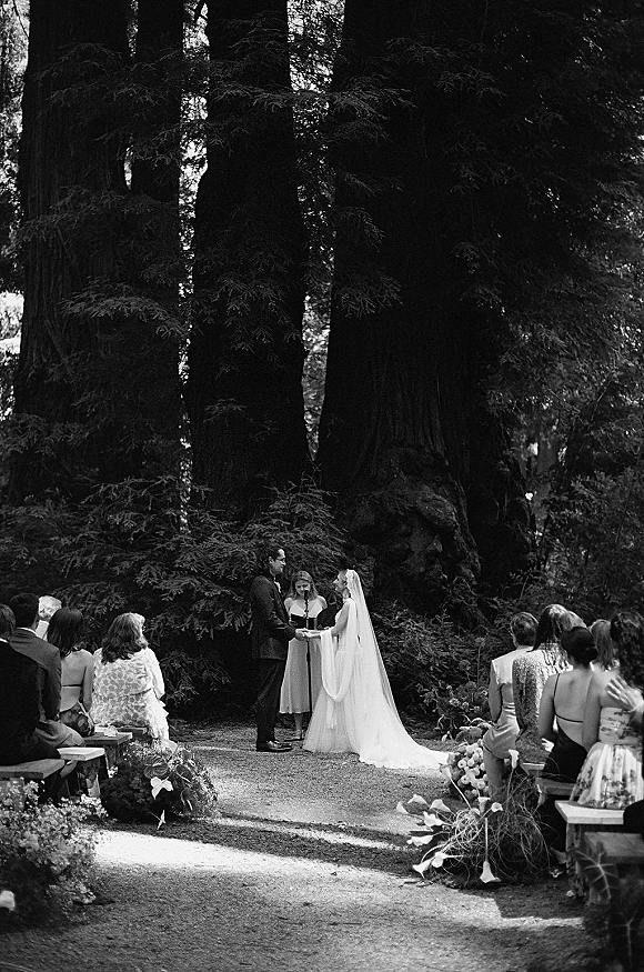 Wedding vows during an outdoor forest wedding ceremony, bride in long veil and groom in dark suit holding hands by floral aisle benches