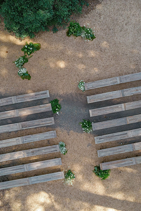 Ceremony setup with outdoor wedding ceremony seating, wood benches lining an aisle with white flowers and greenery in a sunlit forest clearing