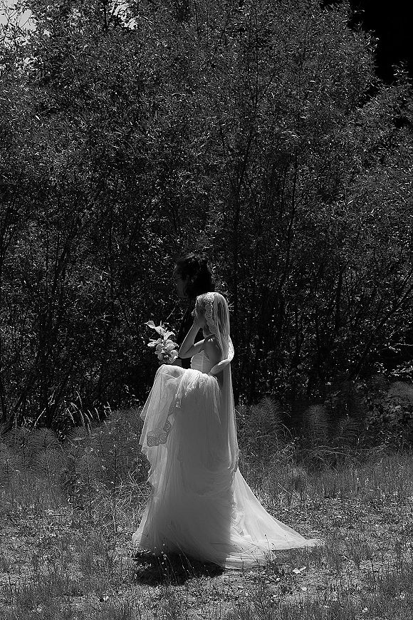 Couple portrait in black and white wedding portrait style, bride in strapless gown with long veil and bouquet embracing groom in a meadow of trees