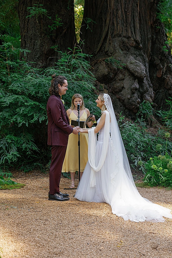 Wedding vows as bride in a cathedral veil and groom in a burgundy suit hold hands while officiant reads in redwood forest