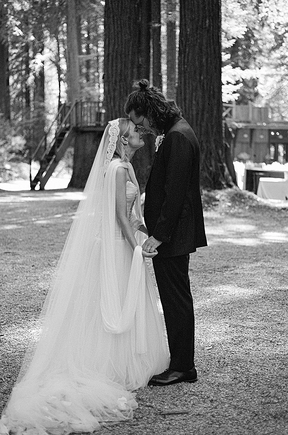 Wedding couple portrait in black and white, bride and groom holding hands with foreheads touching on a forest path, veil trailing behind