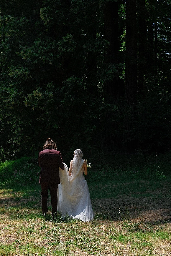 Wedding couple portrait of bride and groom walking away hand in hand through a sunlit forest, bride in veil holding bouquet