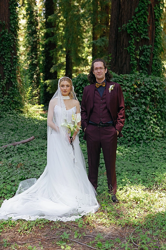 Couple portrait of bride in strapless gown and veil holding calla lilies beside groom in burgundy suit among tall forest trees