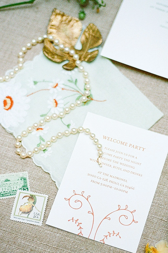 Wedding invitation suite with a welcome party invitation, envelope and vintage stamps, accented by pearl necklace and gold leaf dish on linen