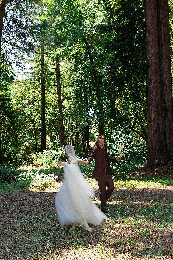 Couple portrait of newlyweds holding hands as the bride twirls her veil and dress, groom in burgundy suit, in a sunlit forest grove