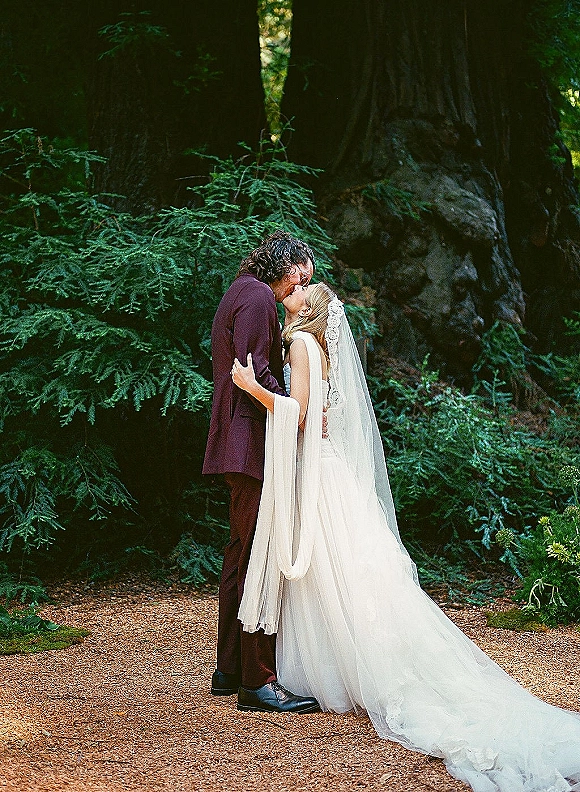 Wedding kiss portrait of bride and groom kissing on a woodland path, her lace veil and dress train flowing beside evergreen trees