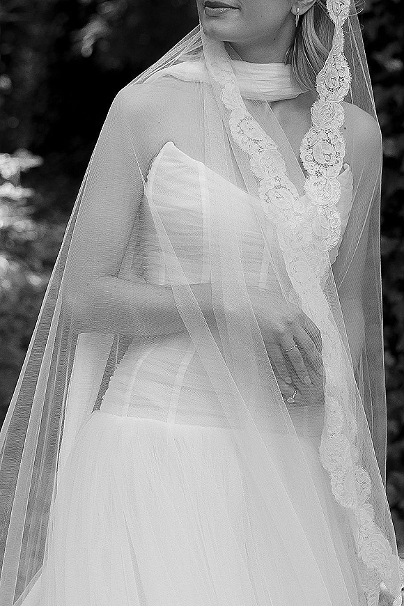 Bridal veil with lace edge draped over shoulders above a strapless wedding dress, drop earrings and ring, set against soft greenery