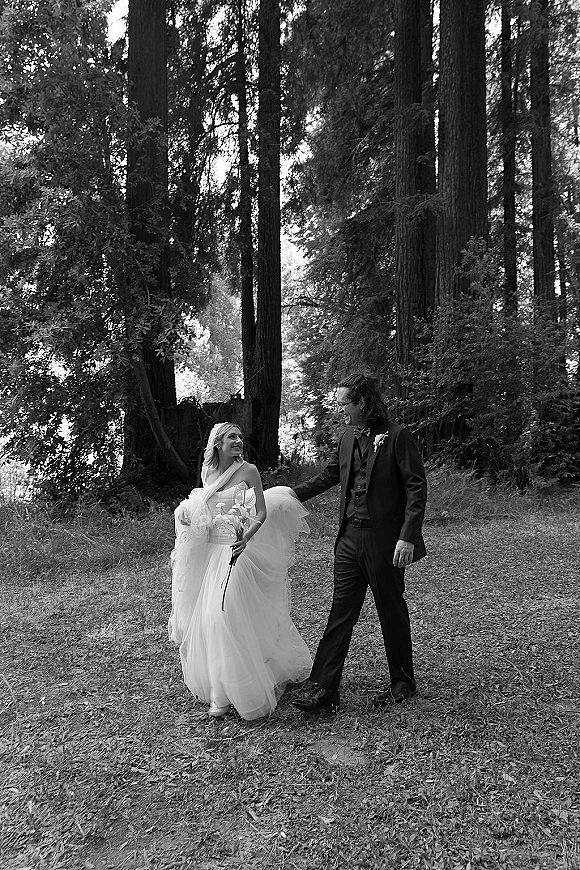 Couple portrait in a black and white wedding photo as bride in strapless dress and veil walks with groom in bow tie through tall forest trees