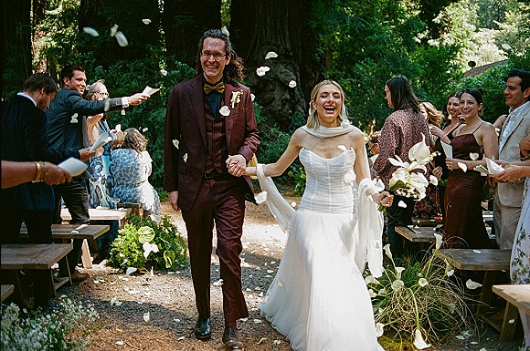 Wedding recessional with bride and groom walking aisle, laughing as guests toss confetti petals along a forest path, veil blowing behind them