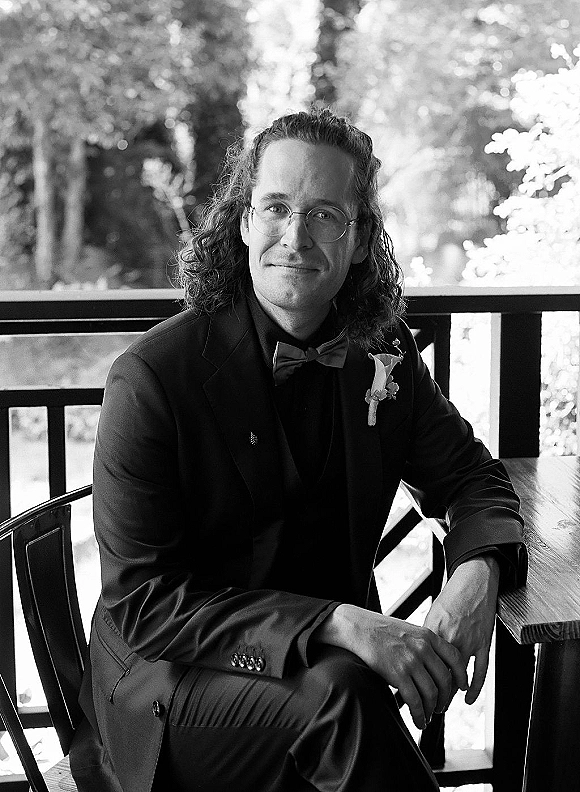Groom portrait in a tuxedo with bow tie, boutonniere, and eyeglasses, seated by a wooden railing with trees behind him