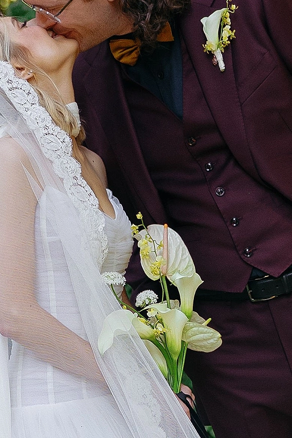 Wedding kiss close up of bride and groom kiss, her lace dress and tulle veil brushing a calla lily bouquet against a dark backdrop