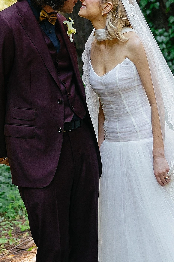 Wedding kiss as bride and groom lean in close, veil and strapless gown visible, groom in suit with calla lily boutonniere amid trees