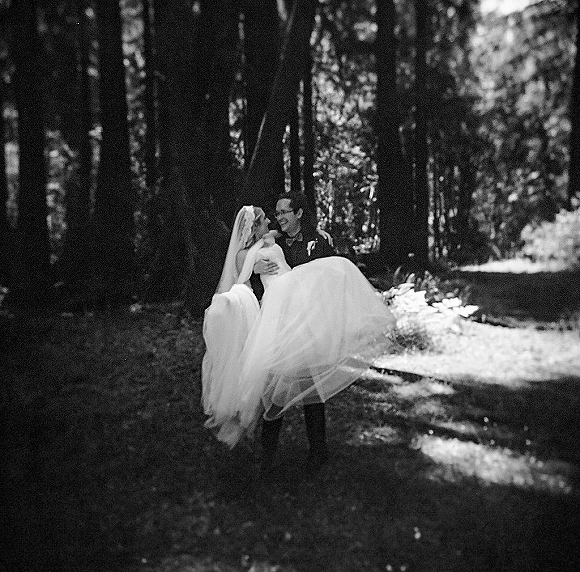 Couple portrait of groom carrying bride in a flowing wedding dress and veil, tuxedo and bow tie, in a forest with dappled sunlight