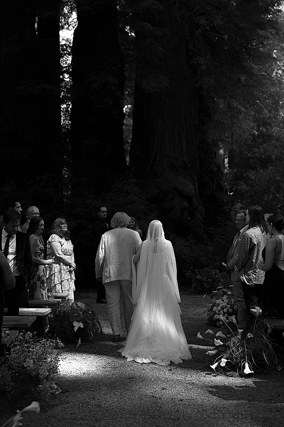 Ceremony processional as bride walking down aisle with a long cathedral veil beside her father on a gravel path under forest trees