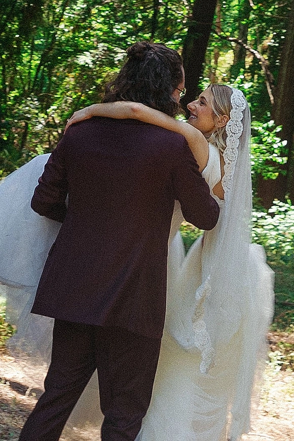 Wedding couple portrait of bride and groom embrace on a sunlit forest path, her white dress and lace veil flowing as they hug