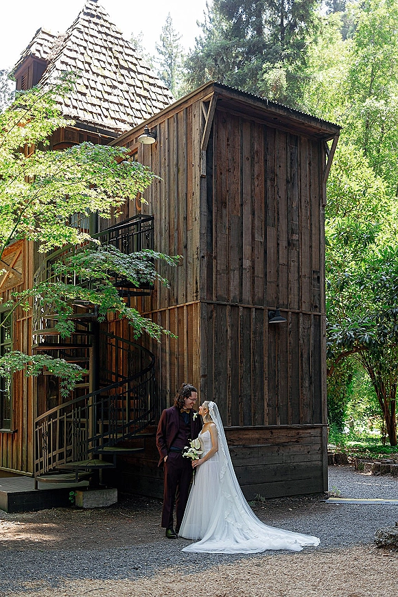 Couple portrait of bride and groom portrait with veil and bouquet, leaning by a wooden building with spiral staircase and trees behind